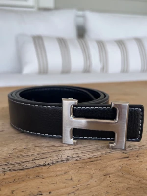 Herme’s bälte med silverfärgat spänne - Snyggt svart skärp i skinn med ett stort, silvrigt H-format metallspänne. Skärpet har vita kontrastsömmar längs kanterna som ger en clean och modern look. Perfekt accessoar för att lyfta vilken outfit som helst. Aldrig använt, sprillans nytt.