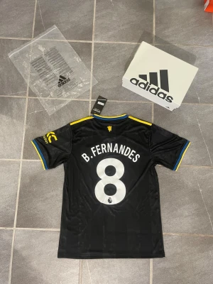 Svart Manchester United fotbollströja - Snygg svart Manchester United fotbollströja från Adidas med gula detaljer och blå inslag på krage och ärmslut. Nummer 8 och B. Fernandes på ryggen. Snapdragon-logga på bröstet och klubbmärke på vänster sida. Perfekt för fotbollsfans!