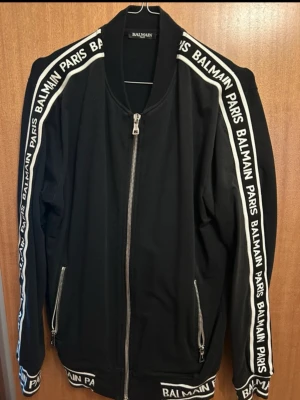 Balmain paris tröja - Snygg svart ziptröja från Balmain Paris med vita logotexter längs ärmar, muddar och nederkant. Tröjan har två framfickor med dragkedja och ribbade detaljer. Perfekt för dig som gillar streetwear och exklusiva märken.