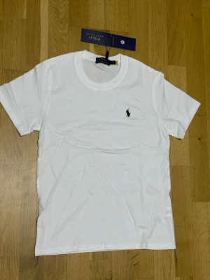 Vit t-shirt från Polo Ralph Lauren - Klassisk vit t-shirt från Polo Ralph Lauren med rund halsringning och korta ärmar. T-shirten har en liten broderad logga på bröstet och är tillverkad i mjuk bomull. Perfekt basplagg för en stilren look.