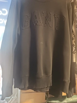 Svart sweatshirt från GANT - Säljer en svart sweatshirt från GANT med stora, upphöjda bokstäver på bröstet. Tröjan har rund halsringning, långa ärmar och ribbade muddar vid ärmslut och nederkant. Perfekt för en avslappnad och stilren look.