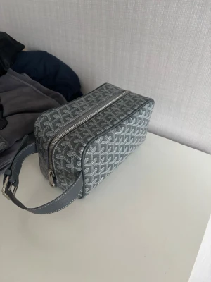 Grå necessär med mönster - Snygg grå necessär med vitt grafiskt mönster och silverfärgad dragkedja. Väskan har ett brett handtag i matchande färg och är tillverkad i ett tåligt material. Perfekt för att organisera dina småsaker eller använda som sminkväska.