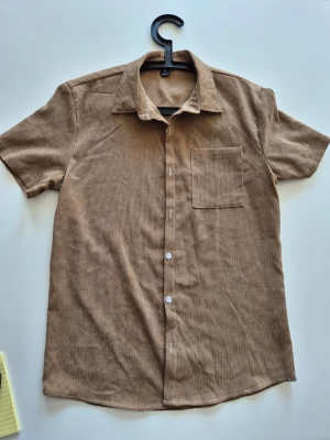 Kortärmad brun manchesterskjorta - Kortärmad skjorta i mjuk brun manchester med klassisk krage och bröstficka. Skjortan har knäppning framtill och en avslappnad passform som passar perfekt till jeans eller chinos.