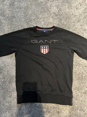 Svart sweatshirt från GANT - Säljer en svart sweatshirt från GANT med broderad logga och ett sköldmärke med stjärnor och ränder på bröstet. Tröjan har rund halsringning och ribbade muddar vid ärmslut och nederkant. Perfekt för en clean och sportig look.