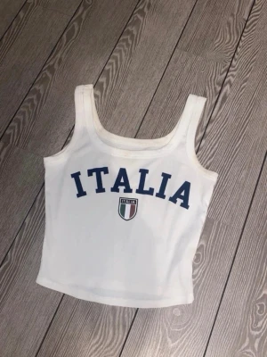 Vit ribbad linnetopp med ITALIA-tryck - Snygg vit ribbad linnetopp med breda axelband och blått ITALIA-tryck framtill. En liten fläck som gör att tvätta bort. 