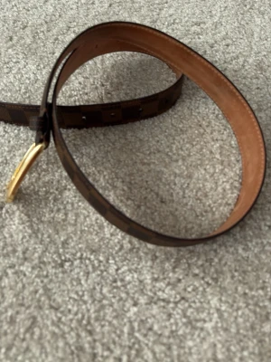 Brunt bälte från louis vuitton  - Snyggt brunt bälte med rutigt mönster och guldfärgat spänne. Bältet har klassisk design och är gjort i skinn med detaljer i mörkbrunt och ljusbrunt. Perfekt accessoar för att lyfta din outfit.