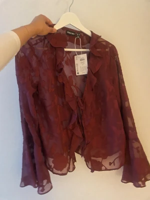 Vinröd transparent blus  - Vinröd blus från Nelly i transparent mesh med blommigt mönster. Blusen har volangdetaljer längs kragen och knytning framtill. Ärmarna är långa och vida, vilket ger en snygg och trendig look. Perfekt för sommaren!! Nypris:300kr