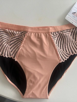 Nya oanvända periodtrosor - Snygga trosor i ljusrosa med dekorativa spetsdetaljerw på sidorna och svarta partier vid grenen. Tillverkade i en mjuk mix av nylon och spandex, med absorberande bomullsfoder och extra lager för komfort. Perfekt för dig som vill ha både stil och funktion.