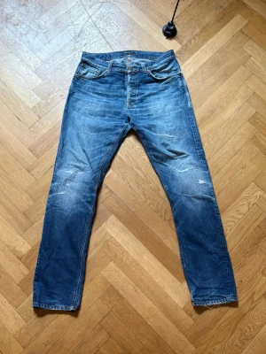 Blå raka jeans med slitningar - Säljer ett par blå jeans med raka ben och snygga slitningar på både fram- och baksida. Jeansen har klassisk femficksdesign och är gjorda i denim. Perfekta för en avslappnad och trendig look.