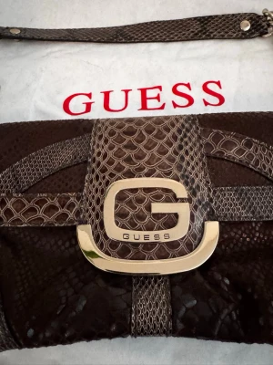 Brun axelremsväska Guess ormmönster - Snygg brun handväska från Guess med ormmönstrad yta och guldfärgat G-spänne framtill. Väskan har axelrem med kedjedetaljer och stängs med lock. Perfekt för dig som gillar statement-accessoarer och trendiga detaljer.