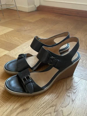 Bekväma kilklackar i skinn - Säljer ett par svarta sandaler med kilklack och två breda remmar i skinn. Båda remmarna har snygga spännen i metall och sulan är i beige gummi med räfflad undersida. Detta är nog de skönaste klackarna jag burit, de är dessutom enkla att styla. Till för dig som vill ha både höjd och komfort i sommarens outfit 🖤
