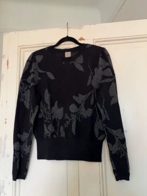 Svart tröja med grått blommönster - Svart finstickad tröja med grått blommönster som ger ett lite glittrigt intryck. Tröjan har långa ärmar och ribbad mudd i midjan och vid ärmsluten. Storlek S. Använd men ändå i ett bra skick! Kontakta om du har frågor💗