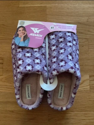 Lila blommiga tofflor från Jomix - Mysiga tofflor från Jomix i lila med vitt och mörklila blommönster. Ovandelen är mjuk och textilklädd, insidan är fluffig och sulan är i ljuslila gummi för bra grepp. Perfekta för att chilla hemma och hålla fötterna varma.