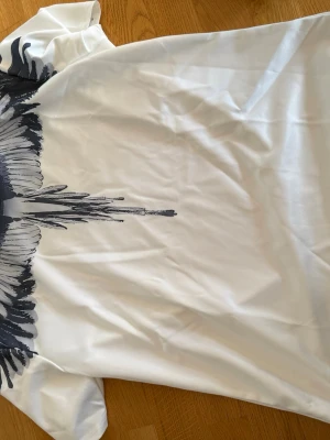Vit t-shirt med svart fjädermönster - Säljer en vit t-shirt med ett stort svart fjädermönster som går över axlarna och ner på bröstet. T-shirten har rund hals och korta ärmar. Materialet känns mjukt och skönt, troligen bomull eller polyester. Perfekt för dig som vill sticka ut lite extra.