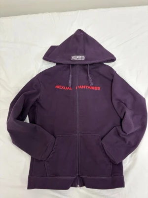 Vetements Sexual Fantasies Hoodie - Lila hoodie från Vetements med dragkedja, huva och fickor framtill. På bröstet står det 'SEXUAL FANTASIES' i rött och på huvan är 'VETEMENTS AUTOMNE-HIVER 2017' broderat i vitt. Tillverkad i mjuk bomull, perfekt för en streetwear-look. Sitter väldigt oversized