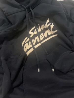 Svart Saint Laurent hoodie - Svart hoodie från Saint Laurent med vit logga tryckt framtill i en cool, handskriven stil. Klassisk huva med dragsko och ribbade muddar vid ärmar och midja. Perfekt för dig som gillar streetwear och vill ha en snygg statementtröja. Tvättlapp och alla andra lappar e avklippta…därför så billigt, självklart äkta dock