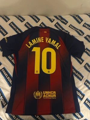 FC Barcelona T-shirt Lamine Yamal - Säljer en officiell FC Barcelona matchtröja med Lamine Yamal och nummer 10 på ryggen. Tröjan är blå och vinröd med gula detaljer, korta ärmar och klubbmärke framtill. Materialet är lätt och andas, perfekt för fotbollsträning eller match.