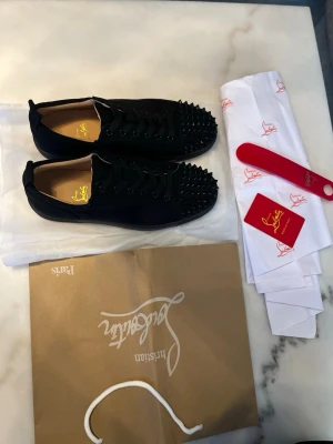 Svarta Louboutin med nitar  - Säljer ett par svarta sneakers från Christian Louboutin med coola svarta nitar på tån och den ikoniska röda sulan. Skorna har snörning och är tillverkade i mocka för en lyxig känsla. Perfekta för dig som vill sticka ut med din stil.