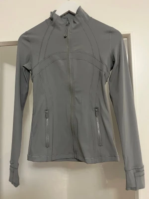 Lululemon Träningsjacka, storlek 4 - Figurnära och mycket smickrande modell med hel dragkedja framtill, hög krage och två fickor med dragkedja. Jackan har flera klassiska Lululemon-detaljer såsom diskret logga bak, fina panelsömmar, stretchigt material och sportig premiumkänsla.  Passar perfekt till yoga, pilates, träning, promenader eller som en snygg vardagsjacka.  Storlek 4, motsvarar XS/S LjusGrå.  Endast testad och redo för ny användare.