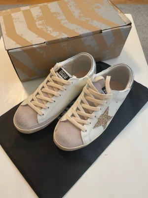 Vita sneakers med glitterstjärna - Snygga vita sneakers från Golden Goose med beige snörning, silverfärgad häl och en glittrig guldstjärna på sidan. Tåpartiet är i ljusgrå mocka och skon har en klassisk låg siluett. Perfekta för dig som vill sticka ut med stilrena detaljer.