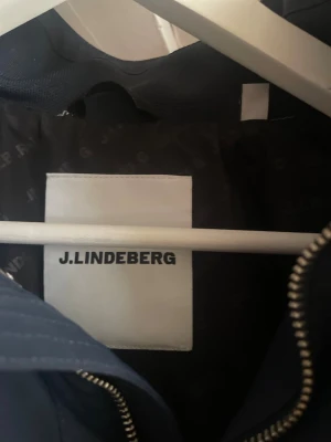 Marinblå vindjacka från J.Lindeberg - Snygg marinblå vindjacka från J.Lindeberg med diskret logga på ärmen och silverfärgade knappar vid kragen. Jackan har dragkedja framtill och är tillverkad i ett lätt och smidigt material, perfekt för blåsiga dagar.