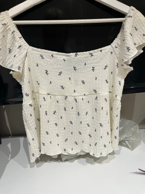 Vit blommig offshoulder topp Divided - Söt offshoulder topp från Divided i vit med små blommiga mönster i svart. Toppen har volangdetaljer vid axlarna, snörning framtill och är tillverkad i ett lätt, krinklat bomullstyg. Perfekt för en fräsch och trendig look.