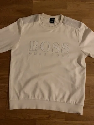 Vit sweatshirt från Hugo Boss - Snygg vit sweatshirt från Hugo Boss med broderad logga på bröstet. Tröjan har rund halsringning, ribbade muddar och kontrastpartier i ljusgrått över axlarna. Perfekt för en clean och stilren look.