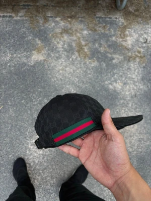 Svart keps med gröna och röda detaljer - Säljer en svart keps med diskret mönster och klassiska gröna och röda ränder på sidan. Kepsen har böjd skärm och justerbar rem bak. Materialet ser ut att vara canvas eller bomull, perfekt för en stilren och trendig look.