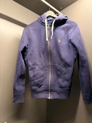 Lila hoodie med dragkedja från Polo Ralph Lauren - Säljer en lila hoodie från Polo Ralph Lauren med dragkedja framtill, vita snören i huvan och den klassiska gula loggan broderad på bröstet. Hoodien har två fickor fram och ribbade muddar vid ärmar och nederkant. Perfekt för en avslappnad stil.