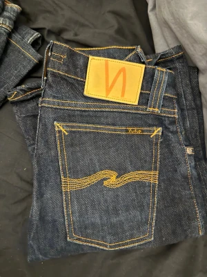 Mörkblå jeans från Nudie Jeans - Säljer ett par mörkblå jeans från Nudie Jeans med klassisk gul kontrastsöm och vågig brodyr på bakfickan. Jeansen har en rak passform och är tillverkade i kraftigt denimtyg. Perfekta för dig som gillar stilrena och tidlösa jeans. Storlek 27/34 slimkim 