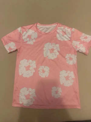 Rosa t-shirt med vita blommor - Säljer en rosa t-shirt i storlek M med vita blommiga mönster. T-shirten har rund halsringning och korta ärmar. Perfekt för dig som vill sticka ut med en färgglad och lekfull stil.