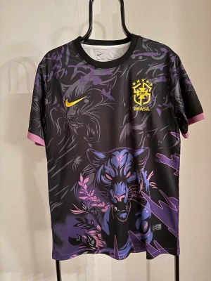 Nike Brasil svart-lila matchtröja - Unik svart och lila matchtröja från Nike med Brasil-logga och gult swoosh. Tröjan har ett stort pantermotiv framtill, grafiska detaljer och klösmärken på ryggen. Korta ärmar med lila muddar och sportig passform. Perfekt för dig som vill sticka ut på planen.