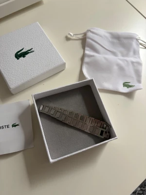 Lacoste Armband - Snyggt armband i silverfärgad metall från Lacoste med rektangulära länkar och diskreta mönster på vissa länkar. Armbandet har en stilren och modern design, perfekt för dig som gillar minimalistiska accessoarer. Levereras i originalask och med tygpåse med Lacoste-logga. Storleken är 21cm men det går att göra det kortare.                                     Skriv om ni har några funderingar!😃