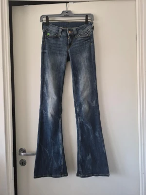 Blå bootcut jeans från Cheap Monday - Säljer ett par blå bootcut jeans från Cheap Monday med slitningar och markerade sömmar. Jeansen har låg midja, klassiska fem fickor och en grön logga bak. Perfekta för dig som gillar retrovibbar och en avslappnad look.
