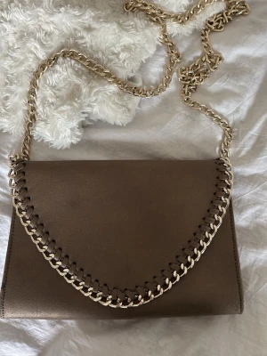 Tiamo - guldig/brun axelväska med guldkedja - Snygg guldig/brun axelväska i mockaimitation med guldfärgad kedja som axelrem och dekorativ kedjedetalj längs locket. Väskan har en stilren, rektangulär form och passar perfekt till en trendig look.