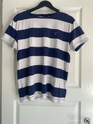 Randig t-shirt från GANT i bomull - Säljer en klassisk t-shirt från GANT med breda marinblå och vita ränder. T-shirten har korta ärmar, rund hals och broderad GANT-logga på bröstet. Materialet är mjuk bomull som känns skön mot huden. Perfekt för dig som gillar stilren och sportig look.