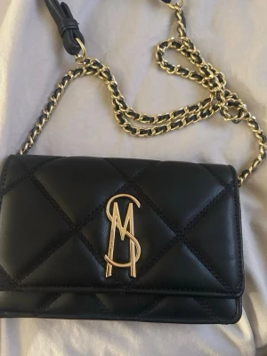 Svart Steve Madden väska - Säljer en Steve Madden väska med guldfärgad kedjerem. Köpte den från boozt för 1079kr men nu säljer jag den för 600❣️har inte använt väskan jätte mycket pga att jag fick en ny, men den är fortfarande i väldigt bra skick. Kontakta via intresse❤️