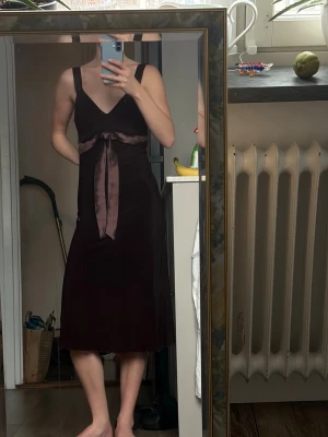 Mörkbrun klänning med satinrosett - Vinröd klänning med breda axelband och djup v-ringning (en storlek för stor på mig. Midjan markeras av ett brett satinband som knyts i en rosett framtill. Fin midiklänning lite carrie bradshaw inspirerande.