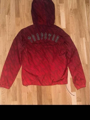 Röd camo vindjacka från Trapstar - Snygg röd vindjacka från Trapstar med camomönster och huva. Jackan har dragkedja framtill, två fickor med knappar och justerbar nederkant. Stor Trapstar-logga på ryggen och liten logga på bröstet. Perfekt för dig som vill sticka ut med streetwear-stil.