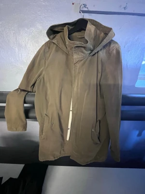 Olivgrön tunn parka från Lager 157 - Säljer en olivgrön parkas från Lager 157 med huva och dragkedja framtill. Jackan har två fickor med knappstängning och en enkel, stilren design. Perfekt för blåsiga vårdagar dagar och lätt att matcha med olika outfits.