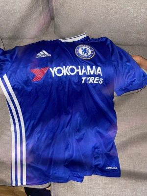 Chelsea FC blå matchtröja Adidas - Chelsea FC matchtröja i blått från Adidas med vita detaljer och Yokohama Tyres-logga på bröstet. Tröjan har korta ärmar, rund hals och klassiska Adidas-ränder längs sidorna. Perfekt för fotboll eller att bära som supporter.