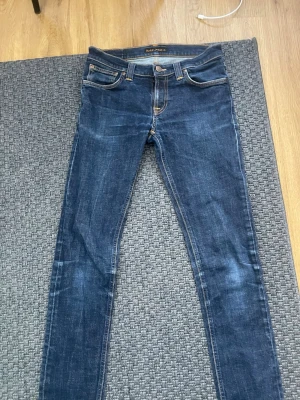 Blå slim fit jeans från Nudie Jeans - Säljer ett par blå slim fit jeans från Nudie Jeans med klassiska fem fickor och snygga kontrastsömmar. Jeansen har en smal passform och är tillverkade i denim med stretch för extra komfort. Perfekt för dig som gillar en modern och stilren look.