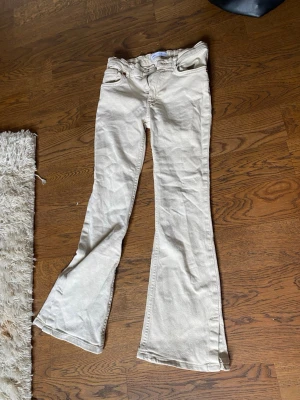 Beiga bootcut jeans med låg midja - Säljer ett par beiga bootcut jeans med låg midja och klassisk femficksdesign. Jeansen har utsvängda ben och är perfekta för dig som gillar en retroinspirerad look. Materialet är denim i bomull och de har bälteshällor samt knappgylf.
