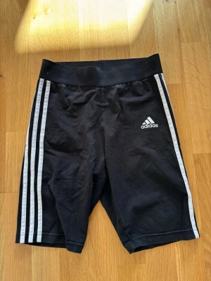 Svarta shorts från Adidas - Snygga svarta cykelshorts från Adidas med klassiska vita ränder längs sidorna och Adidas-logga på låret. Shorts med hög midja och tight passform, perfekta för träning eller sportiga outfits. Tillverkade i stretchigt material för maximal komfort.