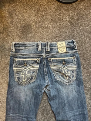 Blå Rock Revival jeans med brodyr - Säljer ett par blå jeans från Rock Revival med raka ben och snygga broderade bakfickor. Jeansen har slitna detaljer och dekorativa knappar på fickorna. Perfekta för dig som gillar en unik och trendig stil. storlek 36, midja 46cm, innerbenslängd 106cm, benöppning 22cm. 