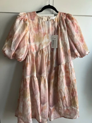 Klänning med puffärm - Supersöt kort klänning med puffärmar och rund hals. Klänningen har ett blommigt mönster i ljusa toner av rosa, beige och gul. Tillverkad i ett lätt och luftigt material, perfekt för varma dagar. Volangdetaljer ger extra volym och en romantisk känsla. Helt ny!! Färgerna är mycket mer ljusa i verkligheten!! Kameran fångar inte färgen så bra. 