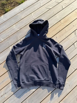 Margaux Dietz Hoodie - Mörkblå hoodie från Brand Store med en liten vit broderad figur på bröstet. Klassisk känguruficka framtill och mjukt material. Perfekt för dig som gillar en enkel och stilren look. Margaux Dietz x Kids Brandstore, 