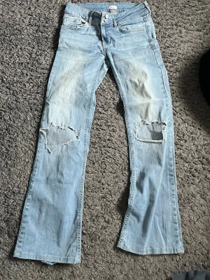 Ljusblå slitna bootcut jeans Nelly - Säljer ett par ljusblå bootcut jeans från Nelly med slitningar och hål på båda knäna. Jeansen har klassisk femficksmodell, låg midja och är tillverkade i denim. Perfekta för en avslappnad och trendig look.