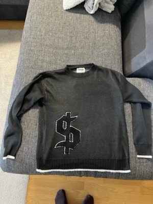 Svart stickad tröja från Buy happiness - Cool svart stickad tröja från buy happiness  med ett stort dollartecken i svart och vitt framtill. Tröjan har rund hals, långa ärmar och ribbade muddar samt vit kant längst ner och vid ärmsluten. Perfekt för dig som gillar streetstyle och statementplagg.