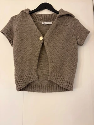 Kortärmad stickad kofta från Zara - Kortärmad stickad kofta från Zara i mjuk beige. Koftan har en stor krage, ribbade kanter och stängs med en rund, ljus knapp framtill. 